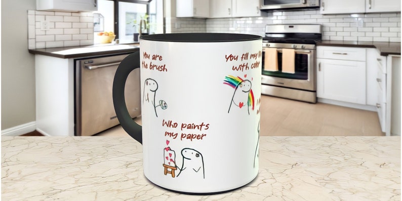 May include: White ceramic mug with a black handle and black interior. The mug has a cartoon illustration of a person with the text "You are the brush", "You fill my life with color", and "Who paints my paper".