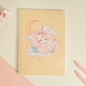 May include: A yellow spiral bound notebook with a pink and white illustration of a fox sleeping in a flower. The illustration is surrounded by clouds and stars. The text "suki" is printed in the bottom right corner.