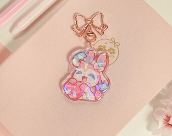 May include: A pink and blue iridescent charm with a cartoon character holding a heart. The charm is attached to a gold-colored bow and a metal ring.