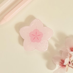 May include: A light pink, five-petal flower-shaped object with a darker pink center and a small floral design. The image also shows a few light pink pencils and a sprig of pink flowers.