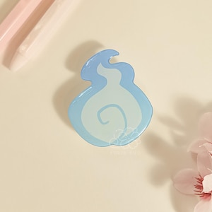 May include: A light blue and white enamel pin in the shape of a flame. The pin has a glossy finish and a swirl design in the center. The pin is on a cream-colored surface with pink pens and flowers.