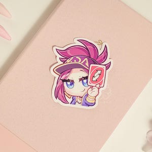May include: A sticker featuring a cartoon character with pink hair, a purple and yellow hat, and a blue and white outfit. The character is holding a red Uno card with the "Draw Two" symbol.