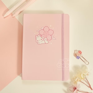 May include: A light pink notebook featuring a white graphic of a small rabbit resting near a cherry blossom flower. The notebook includes a vertical elastic band closure and a faint outline of a flower near the bottom right corner.