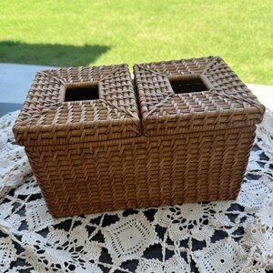 May include: A rectangular, woven wicker basket with two compartments. Each compartment has a square opening in the lid. The basket is a warm brown color and sits on a white, lacy tablecloth.