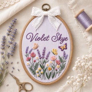 May include: An oval embroidery hoop containing fabric with a light purple and white vertical stripe pattern. The text Violet Skye is embroidered in purple script above a colorful floral scene featuring lavender, daisies, and butterflies.