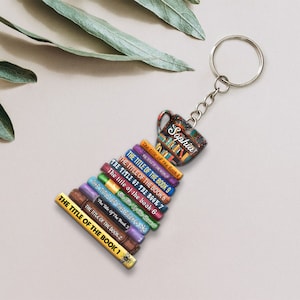 May include: A colorful book stack keychain with a coffee cup charm. The cup has the name "Sophia" written on it. The books are stacked on top of each other and have titles like "The Title of the Book 1" and "The Title of the Book 9".