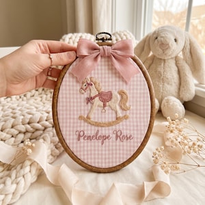 Puede incluir: Un bastidor de bordado ovalado de madera que contiene tela de cuadros vichy en rosa claro y blanco. La tela presenta un caballito balanc&iacute;n bordado en beige y rosa, con el nombre Penelope Rose cosido debajo. Un lazo de cinta rosa empolvado est&aacute; fijado en la parte superior.