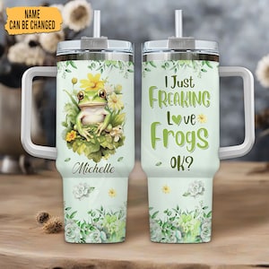 May include: A light green tumbler with a white handle and lid. One side features a frog illustration with the name "Michelle". The other side says "I Just Freaking Love Frogs OK?" in green lettering. Floral and succulent designs decorate the bottom.