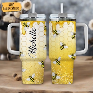 Könnte beinhalten: Ein personalisierter Becher mit Bienenmotiv. Der Becher hat einen weißen Griff und einen silbernen Deckel mit Strohhalm. Das Design umfasst ein Wabenmuster, tropfenden Honig und Illustrationen von Bienen. Der Name "Michelle" ist in einer Schreibschrift geschrieben.