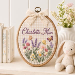 May include: An oval wooden embroidery hoop containing fabric with a beige and white gingham pattern. The fabric features hand-stitched purple text reading Charlotte Mae, surrounded by embroidered lavender, pink flowers, and three butterflies.