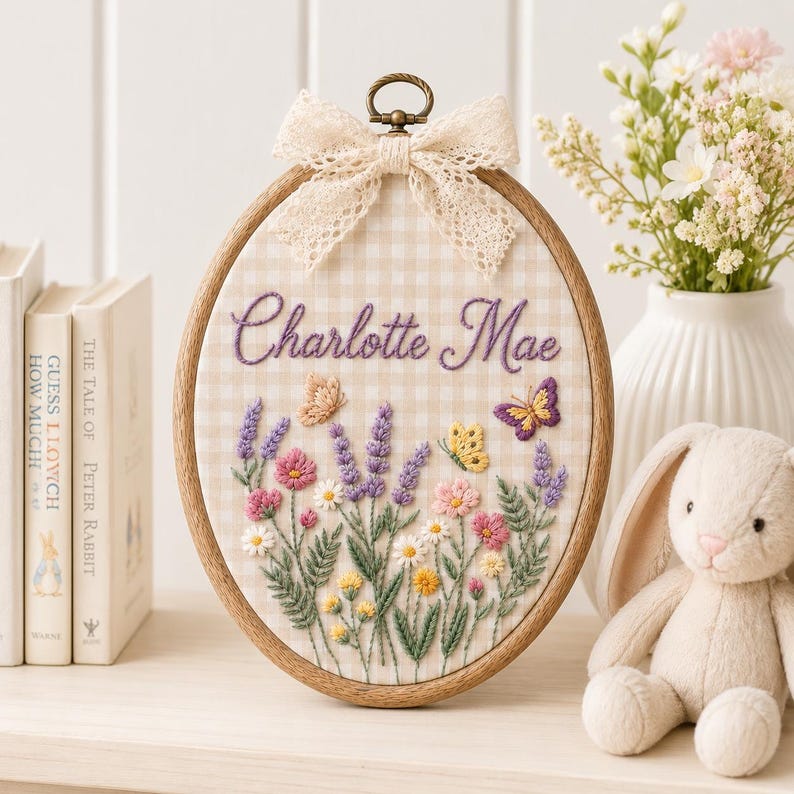 May include: An oval wooden embroidery hoop containing fabric with a beige and white gingham pattern. The fabric features hand-stitched purple text reading Charlotte Mae, surrounded by embroidered lavender, pink flowers, and three butterflies.