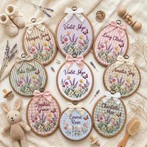 May include: A collection of nine circular wooden embroidery hoops featuring personalized names and floral designs with lavender, wildflowers, and butterflies. Each hoop is topped with a decorative fabric bow and displays a unique gingham or striped background.