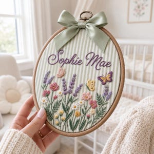 May include: An oval wooden embroidery hoop containing fabric with green and white vertical stripes. The fabric features hand-stitched floral designs, butterflies, and the name Sophie Mae in purple script. A sage green satin ribbon bow is attached.