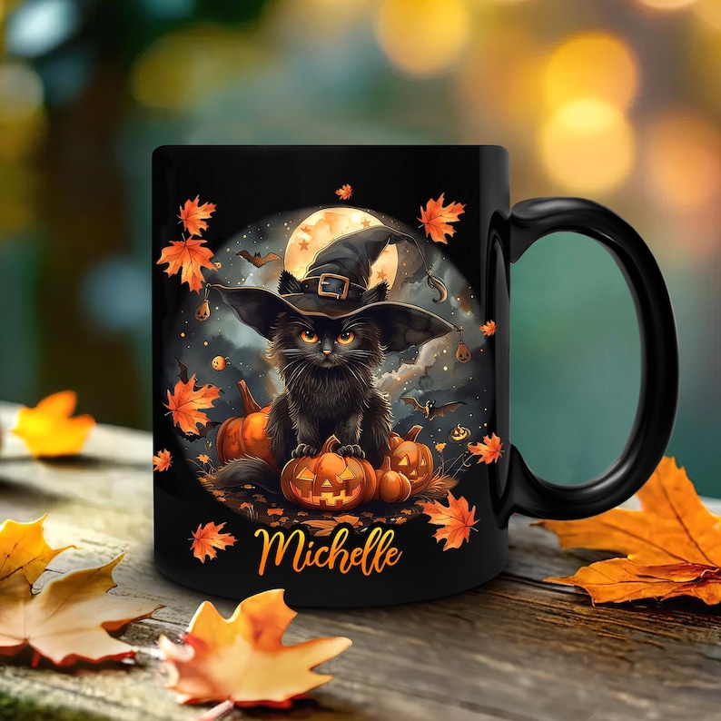 Op de afbeelding: Zwarte keramische mok met een Halloween-ontwerp. Het toont een zwarte kat met een heksenhoed, pompoenen, een volle maan en herfstbladeren. De naam "Michelle" staat op de mok.