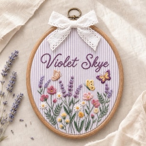May include: An oval wooden embroidery hoop containing fabric with a light purple and white vertical stripe pattern. The embroidery features the name Violet Skye in purple script, surrounded by lavender sprigs, wildflowers, and butterflies.