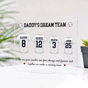 May include: White acrylic sign with a baseball theme. It says "Daddy's Dream Team" with four baseball jerseys with names and numbers. The text below says "We are your number one fans always and forever and together we make a winning team."