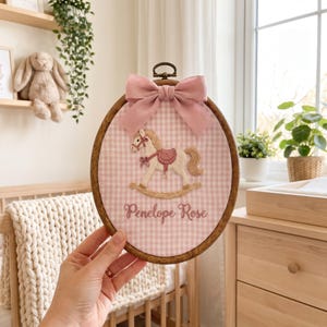 Puede incluir: Un bastidor de bordado ovalado de madera con tela de cuadros vichy rosa y blanca. La tela presenta un caballito balanc&iacute;n bordado en beige y rosa, con el nombre Penelope Rose cosido debajo en hilo rosa. Un lazo de tela rosa empolvado est&aacute; fijado.