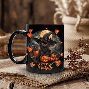 Op de afbeelding: Zwarte keramische mok met een Halloween-ontwerp. Het ontwerp toont een zwarte kat met een heksenhoed, pompoenen, een volle maan en herfstbladeren. De naam "Michelle" staat op de mok.