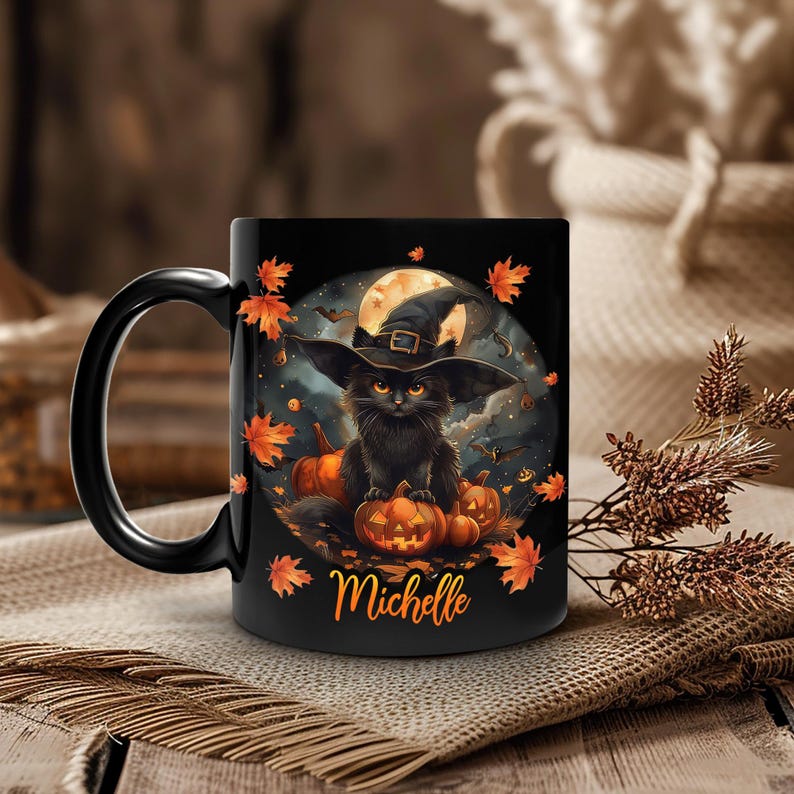 Op de afbeelding: Zwarte keramische mok met een Halloween-ontwerp. Het ontwerp toont een zwarte kat met een heksenhoed, pompoenen, een volle maan en herfstbladeren. De naam "Michelle" staat op de mok.