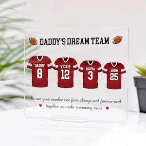 May include: A red and white acrylic sign with the text "Daddy's Dream Team" and four red and white football jerseys with the names "Daddy", "Vickie", "Silvia", and "John" and the numbers 8, 12, 3, and 25. The text below the jerseys reads "We are your number one fans always and forever and together we make a winning team."