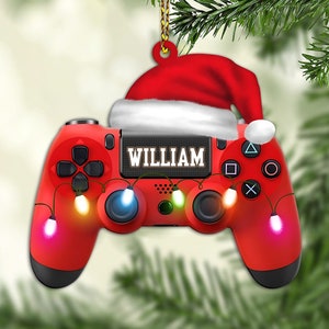May include: Red and black video game controller Christmas ornament with a Santa hat on top. The controller has a string of colorful lights wrapped around it and the name "WILLIAM" is printed on the front.