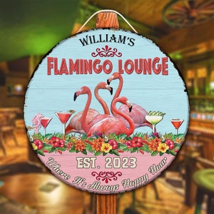 May include: A round wooden sign with a blue background and pink flamingos. The sign says "William's Flamingo Lounge Est. 2023 Where It's Always Happy Hour".