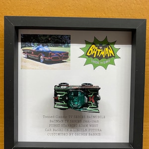 Puede incluir: Una vitrina enmarcada que presenta una réplica de coche de juguete negro y verde del Batimóvil de la serie de televisión Batman de la década de 1960. La vitrina incluye una foto del Batimóvil original y un logotipo vintage de Batman. El texto dice: "Tooned Classic TV Series Batmobile, Batman TV Series 1966-1968, First Starring Adam West, Car Based on a Lincoln Futura, Customized by George Barris."