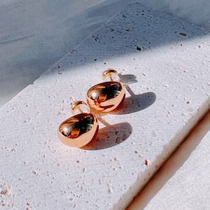 May include: A pair of gold-toned stud earrings with a smooth, rounded shape. The earrings are on a white textured surface.