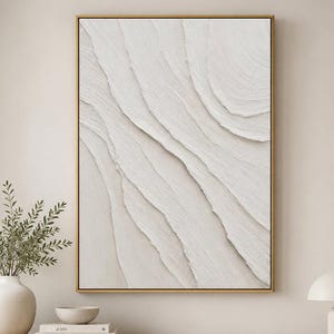 Puede incluir: Una pieza de arte mural rectangular que presenta un diseño abstracto texturizado en color blanco roto con crestas onduladas superpuestas. La obra está enmarcada en un fino marco de metal dorado y se muestra verticalmente, con una paleta de colores neutros.