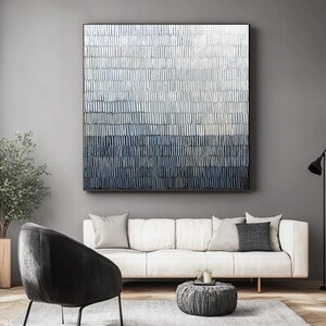 May include: Large abstract wall art in shades of blue, grey, and white. The artwork features a textured, layered design with vertical brushstrokes. The piece is framed in black and hangs above a white sofa in a modern living room.