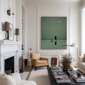 May include: A framed art print depicts a person walking a dog on a white line against a green background. The artwork has a minimalist style, with a reflection of the person and dog below. The room features a fireplace, armchair, and coffee table.