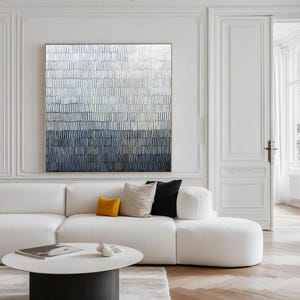 May include: Abstract wall art with a textured design in blue, grey, and white tones. The artwork is framed and hangs above a white sofa with cushions. A round coffee table and white walls complete the modern interior design. The artwork is a modern home decor piece.