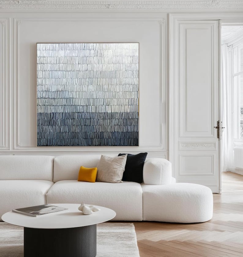 May include: Abstract wall art with a textured design in blue, grey, and white tones. The artwork is framed and hangs above a white sofa with cushions. A round coffee table and white walls complete the modern interior design. The artwork is a modern home decor piece.