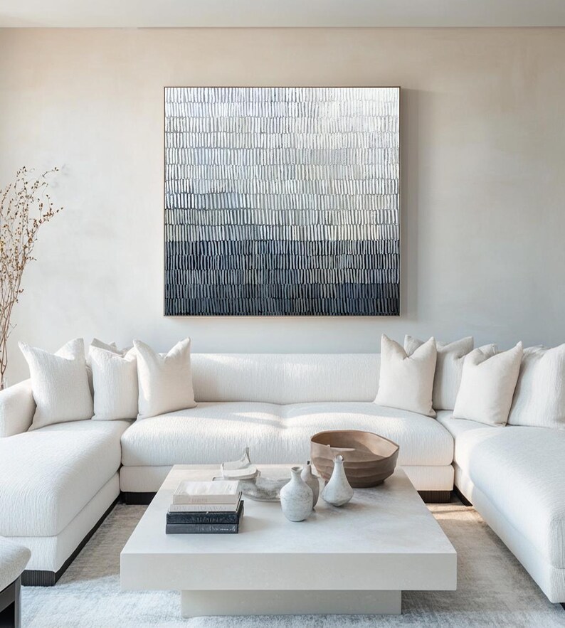 May include: A contemporary living room with an abstract artwork in shades of white, grey, and navy blue. A white corner sofa, a white square coffee table, and decorative items such as vases and a wooden bowl are also visible.