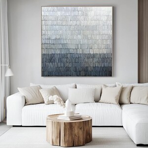 May include: An abstract painting with a textured surface, displaying a colour gradient from light grey to dark blue. It hangs above a white sectional sofa with beige cushions, and a wooden stump coffee table.
