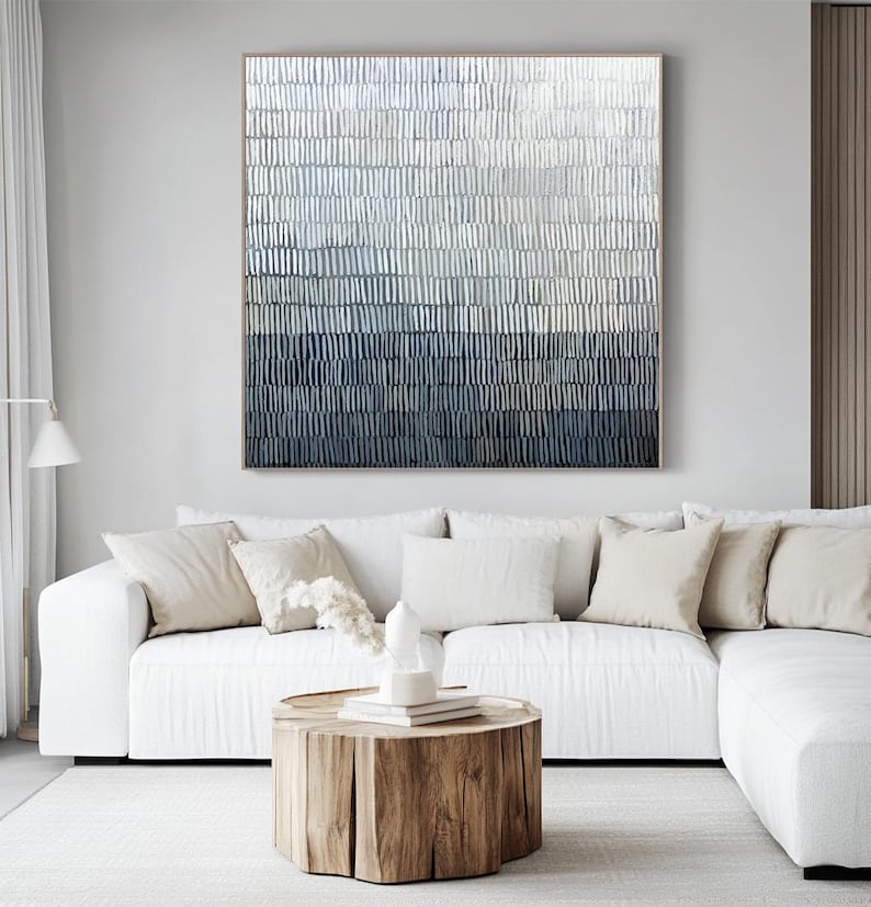 May include: An abstract painting with a textured surface, displaying a colour gradient from light grey to dark blue. It hangs above a white sectional sofa with beige cushions, and a wooden stump coffee table.