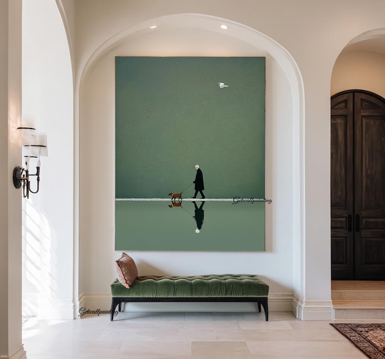 May include: A large vertical canvas print depicts a person walking a dog along a white line, reflected in the water below. The artwork features a muted green and white color palette, with a single bird in the sky. A green velvet bench sits below the artwork.