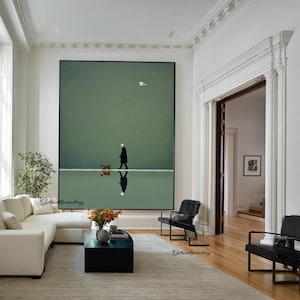 May include: A large framed art print depicts a person walking a dog on a reflective surface, with a bird in the sky. The artwork is set against a muted green background, creating a serene and minimalist aesthetic. The scene is reflected in the water.