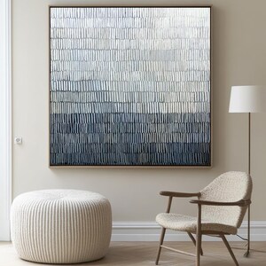 May include: Abstract wall art in a wooden frame. The artwork features a gradient of vertical brushstrokes in shades of white, grey, and navy blue. The room also includes a beige ottoman, a wooden chair, and a floor lamp.