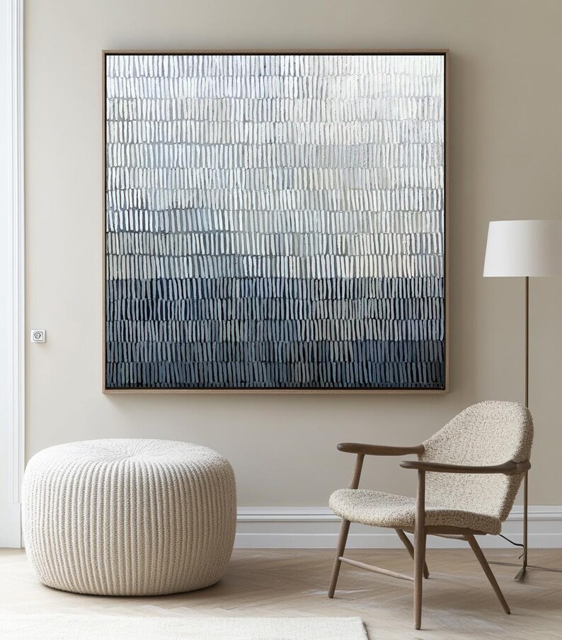 May include: Abstract wall art in a wooden frame. The artwork features a gradient of vertical brushstrokes in shades of white, grey, and navy blue. The room also includes a beige ottoman, a wooden chair, and a floor lamp.