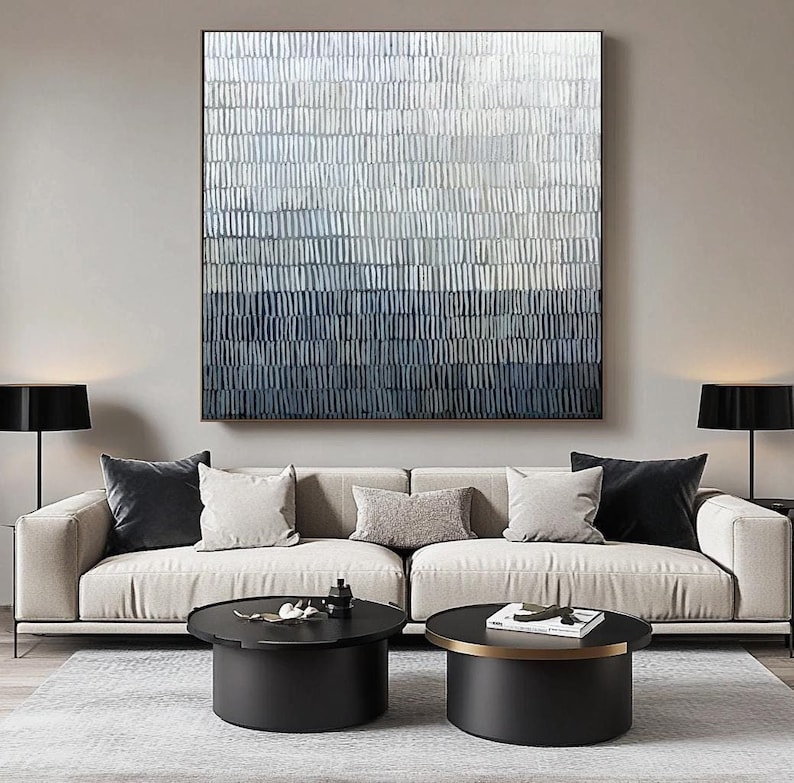 May include: Abstract wall art with a textured, gradient design in shades of white, blue, and navy. The artwork is framed and hangs above a beige sofa with decorative cushions. Two black coffee tables and lamps complete the modern living room setting.