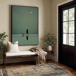May include: A framed art print depicts a person walking a dog on a reflective surface, with a bird in the sky. The artwork is set against a muted green backdrop, creating a serene and minimalist aesthetic. The print is displayed above a bench with a cream-colored cushion.
