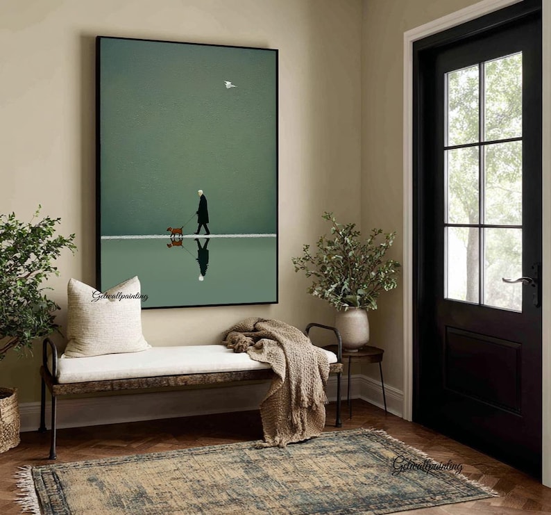 May include: A framed art print depicts a person walking a dog on a reflective surface, with a bird in the sky. The artwork is set against a muted green backdrop, creating a serene and minimalist aesthetic. The print is displayed above a bench with a cream-colored cushion.