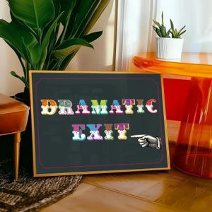 May include: A black framed sign with a gold border. The sign has the words "Dramatic Exit" written in colourful, stylized letters. A hand with a pointing finger is pointing to the right.