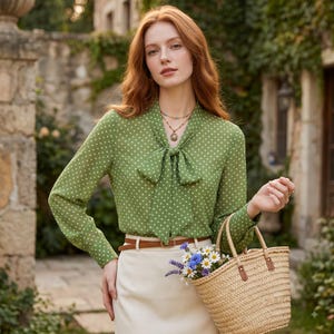 Op de afbeelding: Een groen shirt met lange mouwen, een wit stippenpatroon en een strikkraag, gecombineerd met een crèmekleurige rok met een bruine riem en een geweven rieten mandtas met een klein boeket witte en blauwe bloemen.