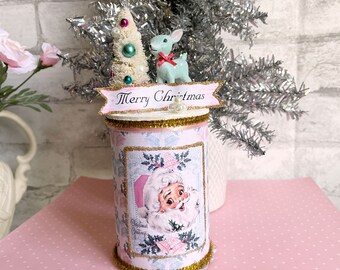 Puede incluir: Un contenedor cilíndrico de Navidad rosa y blanco con una imagen de Papá Noel, un pequeño árbol de Navidad blanco con adornos rosas y verdes, y una figurita de reno turquesa. El contenedor tiene un borde de purpurina dorada y una pancarta que dice "Feliz Navidad".