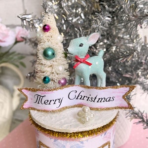 K&ouml;nnte beinhalten: Eine wei&szlig;e Flasche mit rosa und goldenen Verzierungen, verziert mit einem kleinen wei&szlig;en Weihnachtsbaum mit bunten Ornamenten, einer blauen Hirschfigur und einem rosa Banner mit der Aufschrift "Frohe Weihnachten".