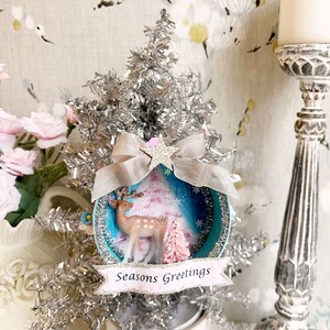 Op de afbeelding: Een blauwe en witte kerstbal met een zilveren tinselboom, een roze en witte boom, een hertenfiguur en een banner met de tekst "Seasons Greetings".
