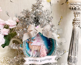 Puede incluir: Un adorno navideño azul y blanco con un árbol de oropel plateado, un árbol rosa y blanco, una figura de ciervo y una pancarta que dice "Seasons Greetings".