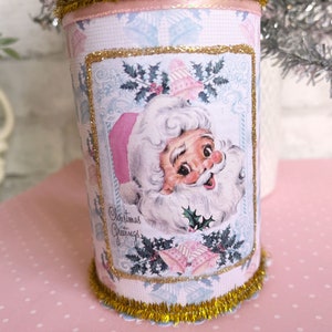 K&ouml;nnte beinhalten: Eine rosafarbene und blaue Weihnachtsdose mit goldener Glitzerverzierung. Die Dose zeigt eine Vintage-Illustration des Weihnachtsmanns mit dem Text "Christmas Greetings".
